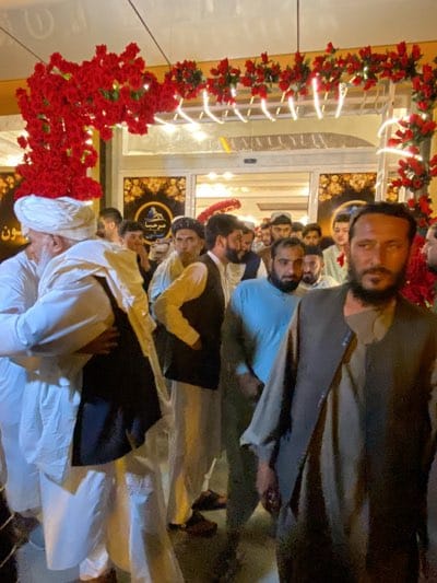 The image depicts a gathering of men, primarily wearing traditional clothing, outside a building entrance adorned with red floral decorations. The scene appears to be located in قندهار, Afghanistan, based on the signage visible on the building. The men are diverse in age and appearance, with some wearing turbans and traditional vests. The overall impression is that of a formal event or gathering taking place at the location.