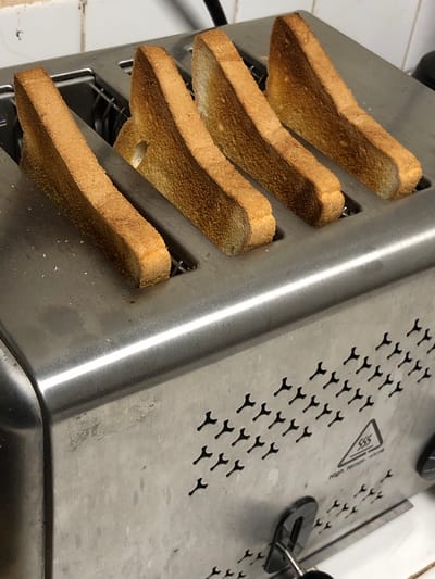 A stainless steel toaster is shown with four slices of toasted bread in its slots. The bread slices are evenly browned and appear to be standard white bread. The toaster has a modern design with a brushed metal finish. There are ventilation holes on the side of the toaster, forming a decorative pattern. Below the ventilation holes, there is a warning symbol indicating "High temperature". The toaster is placed on a white tiled surface, likely a kitchen countertop. The background shows white tiled walls. The lighting is bright and consistent, suggesting an indoor environment. There is no indication of the location being Kabalana, Sri Lanka, as this is an indoor household appliance scene.