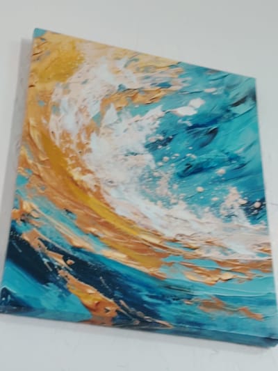 An abstract painting of waves with thick impasto brushstrokes is displayed on a white wall. The painting features vibrant blues, turquoises, golden yellows, and white, creating a dynamic and textured representation of water. The artwork is hung on a plain white wall, likely in a residential or gallery setting within the city of Toranghylyk, Kazakhstan. No people are present in the image, and the focus is solely on the artwork.