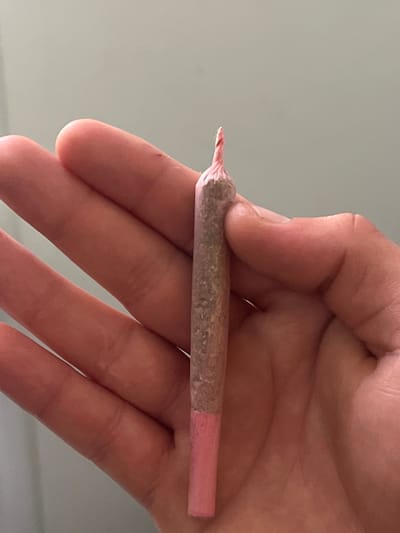 The image shows a person's hand holding a joint. The joint is rolled with pink paper and contains ground plant material. The background is a plain, light-colored wall. There is no information to suggest this image is located in Neuss, Germany.