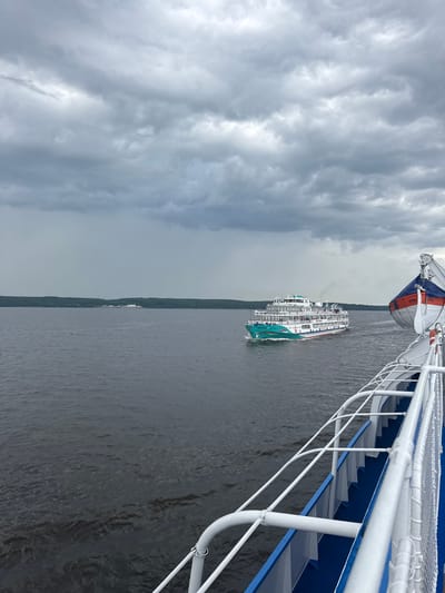 A wide, dark gray river dominates the lower half of the image, with ripples indicating a moderate current. On the right side of the frame, the white deck of a vessel is visible, featuring blue trim and white railings. A life raft, orange and white, is mounted on the side of this vessel. In the distance, a white and teal passenger ship with multiple decks sails on the river, emitting a thin plume of smoke. The far bank of the river is lined with a dense, dark green forest. The sky above is overcast with heavy, gray clouds, suggesting a potentially stormy or rainy day. The scene appears to be set in Ikskoe Ust'e, Russia, given the presence of riverine and forested landscapes characteristic of the region.