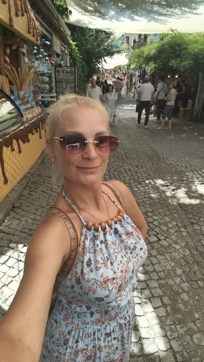 A blonde woman with sunglasses and a floral dress smiles at the camera in a selfie taken on a cobblestone street in Selçuk, Turkey. In the background, other people walk down the street, which is lined with shops and trees. The woman is wearing a light blue floral dress with a halter neckline and wooden beaded accents. She has a brown crossbody bag slung over her shoulder. The street is paved with gray cobblestones and is partially covered by a light-colored awning, casting shadows on the ground. To the left of the woman, a large, yellow ice cream cone-shaped display is visible, with shelves of ice cream cones behind it. The overall scene suggests a tourist area with shops and food vendors.