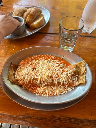 A high-angle shot captures a meal arrangement on a wooden table, likely in a restaurant setting. A plate of pasta topped with tomato sauce and shredded cheese takes center stage. Next to it, a glass of water and a breadbasket containing several slices of bread, along with a small metal ramekin of dip, is also visible. A hand is reaching towards the breadbasket. The location of the scene is Villa General Belgrano, Argentina.