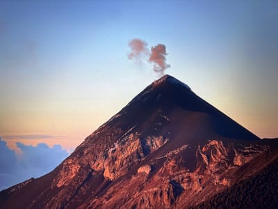 The image depicts a large, conical volcano emitting smoke from its peak. The volcano's slopes are varied in texture, with areas of dark rock, reddish-brown, and areas that appear to have sparse vegetation. The sky behind the volcano is a gradient of light blue to pale orange, suggesting dawn or dusk. Clouds are visible on the left side of the frame. The image does not appear to have anything to do with Placencia, Belize.