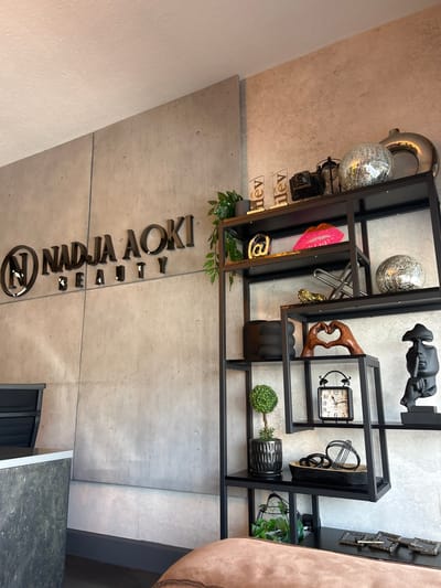 This media file showcases a modern interior space, likely a salon or spa, given the "NADJA AOKI BEAUTY" signage prominently displayed on a concrete wall. The decor features a black metal shelving unit filled with various decorative items. Among these objects are a disco ball, a pink lip-shaped novelty item, a small golden statue, a decorative wooden hand sculpture, a framed clock with Roman numerals, and several black decorative spheres. Green plants in pots add a touch of nature to the display. The lighting appears to be artificial, suggesting an indoor setting. The overall aesthetic is sleek and contemporary, with a focus on metallic and concrete textures. The angle of the photograph is unusual, appearing to be taken from a low vantage point and rotated, which might give the impression of the objects floating or being viewed from an unconventional perspective. There are no people visible in the frame, and the setting doesn't provide clear cues for the time of day or weather.