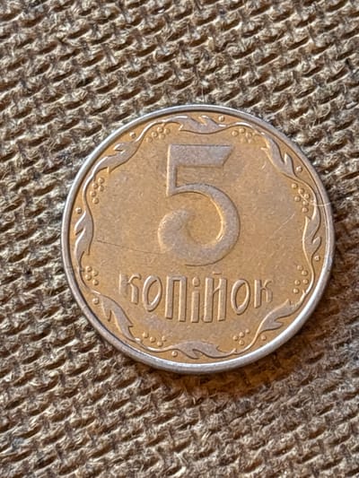 The image shows a close-up, overhead view of a single Ukrainian 5 kopiyok coin resting on a textured, woven surface, likely burlap or a similar coarse fabric. The coin is golden-yellow and circular, with a decorative border featuring a pattern of leaves and small circles. Dominating the center of the coin is a large, stylized number "5". To the right of the "5", the word "копійок" is inscribed in Cyrillic script, indicating the denomination. The lighting suggests an indoor environment, with the sunlight casting a subtle shadow to the lower right of the coin. There are no people or other objects visible in the frame. The background fabric is a warm brown color, with a rough, woven texture that creates a repeating pattern of raised threads. The image appears to be taken in natural light, possibly during the daytime, given the overall brightness and absence of artificial shadows. The location context is Pereiaslavske, Ukraine, which is consistent with the coin's origin.