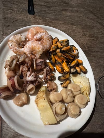 This media file shows a close-up, overhead shot of a white plate filled with various seafood and vegetables, placed on a rustic wooden surface. The plate is full, with portions arranged in a somewhat haphazard manner.

On the upper left side of the plate are a cluster of cooked mussels, their shells a mix of dark blue and orange. Next to them, towards the top and center, are several cooked button mushrooms, their caps a pale beige. Partially obscured by the mushrooms is a piece of cooked artichoke, its layered texture visible.

In the bottom left quadrant of the plate are several cooked shrimp, their shells a translucent pinkish-orange. Adjacent to the shrimp, occupying the bottom center and right, is a generous portion of cooked calamari, cut into rings and tentacles, displaying a reddish-brown hue. There are also a few scattered pieces of what appear to be cooked garlic or another vegetable, particularly on the right side of the plate.

The overall impression is of a meal, possibly an appetizer or a side dish, being served. The lighting is warm, suggesting an indoor setting, and the wooden surface could be a table. There is no visible indication of the time of day or weather. No text is visible in the image. The context provided places this scene in Carpignano Salentino, Italy.