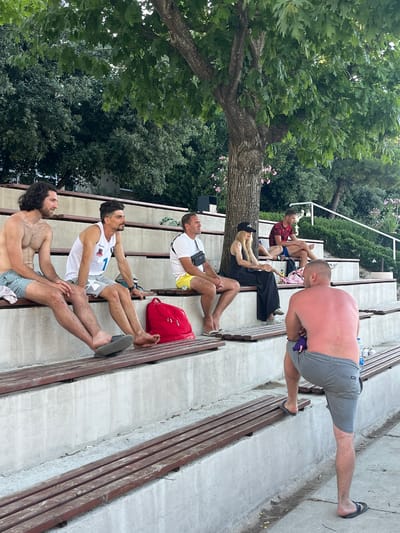 The image depicts an outdoor scene in Gošići, Montenegro, with a group of people seated on tiered concrete benches. In the foreground, a shirtless man with sunburnt skin and gray shorts stands facing away from the camera, with his left leg on a bench. Behind him, several individuals are seated on the benches. From left to right, a man with long dark curly hair and patterned swim trunks is seated. Next to him is a man in a white tank top with a number "1" and gray shorts. To his right, a man in a white t-shirt and yellow shorts is seated. Further back, a woman in a black outfit and cap is seated, and next to her, a man in a red and black patterned shirt and blue shorts is seated. A red backpack is visible on the bench in front of the man in the white tank top. The environment includes lush green trees overhead and a slightly overcast sky.