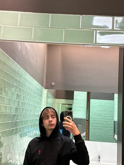 A person in a black hoodie takes a mirror selfie in a bathroom. The bathroom has mint green tiles on the walls and floor, gray walls, and a modern design. The person is holding their phone with a ring light attached. Their reflection shows them looking at the camera. The image is rotated, with the top of the photo being the left side of the bathroom.