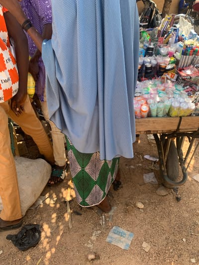The image depicts an outdoor market scene in Boi A, Nigeria. The foreground is dominated by the legs and lower torsos of several people, suggesting a close-up perspective of individuals engaged in or observing the market activity. One person is wearing a light blue and white striped garment, partially covering their lower body. Another is dressed in a purple patterned fabric. A third person is wearing tan pants. The ground is sandy and littered with debris, including plastic bags and some dry vegetation.

In the background, a cart is laden with an assortment of items, including what appear to be flashlights or small lamps packaged in plastic, pens, and various other goods. A bicycle wheel and part of its frame are also visible, suggesting that the cart might be attached to or near a bicycle. The lighting and shadows indicate it is daytime, likely during the morning or afternoon. The overall impression is one of a bustling, informal marketplace, characteristic of many communities in Nigeria. The arrangement of goods on the cart implies that a sale or trade is in progress, or about to commence.