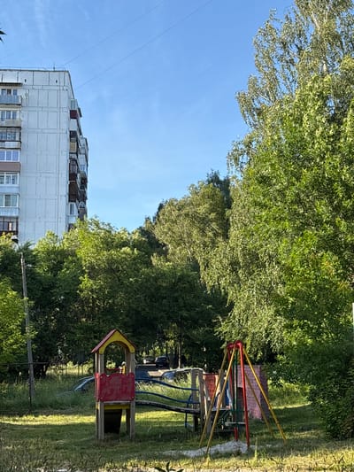 The image depicts an outdoor scene with a clear blue sky and lush green trees. In the foreground, a children's playground is visible with colorful equipment, including a merry-go-round with red, yellow, and green poles, and a small red and yellow playhouse. The playground is situated on a grassy area with some weeds. Behind the playground, a large apartment building with a light-colored facade is prominent. The building has many windows and balconies, suggesting a residential area. The image is captured during the daytime, likely a sunny day, given the bright lighting and clear sky. There are no people visible in the image, and no text is present. The scene appears to be a typical urban or suburban setting, possibly in Sarov, Russia, as indicated. The overall mood is peaceful and sunny.