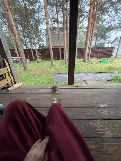 A person is sitting on a wooden porch, seen from above and to the side. They are wearing maroon sweatpants and a white sneaker. Their hands are resting on their lap, with one hand wearing a ring. A small, colorful spinning toy is on the wooden planks near their feet. The porch overlooks a grassy area with trees, a brown wooden fence, and a house in the background. The weather appears overcast. The overall scene suggests a casual, outdoor setting, possibly in a rural or suburban area.