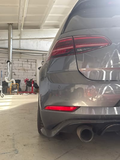 The image shows the rear of a dark gray Volkswagen Golf parked in a garage. The car's taillight, bumper, and exhaust pipe are visible. The garage has a concrete floor and a white ceiling with exposed beams. In the background, there are tools and equipment, including a red engine stand and a yellow fan. Reflections on the car's paint reveal other vehicles, ladders, and a person crouching. The scene is likely in Schkopau, Germany, based on the context of the original image source.