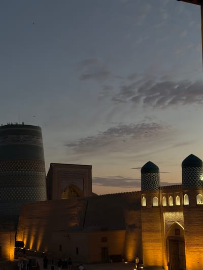 This is a photograph of the city of Khiva, Uzbekistan, at dusk. In the foreground, there are several buildings with brick walls, illuminated by warm, artificial lights. One of these buildings features a large wooden gate. To the right, two cylindrical structures topped with conical green domes rise from behind a wall. These domes are decorated with intricate geometric patterns. In the middle ground, a long, imposing brick wall stretches across the scene, with a large arched entrance visible within it. To the left, a tall, cylindrical tower with a distinctive patterned surface and a decorative band of Arabic script dominates the left side of the frame. Atop this tower, several figures are visible, along with some antenna-like structures. The sky above is a gradient of blues and purples, with scattered clouds tinged pink from the setting sun. A few small birds are captured in flight against the twilight sky. In the lower left corner, a street scene unfolds with a few pedestrians and market stalls, also lit by artificial lights, creating elongated shadows.