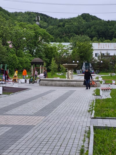 In Uglegorsk, Russia, a paved park area features a prominent fountain with sculpted figures in its center. A man in a black sweater and blue jeans walks past the fountain, carrying a black plastic bag in his left hand and walking his orange bicycle with his right. To his left, the fountain is surrounded by lush green grass and white benches with decorative bases. In the background, a playground with slides and swings is visible, with several people, including a woman in a yellow jacket and a man in an orange jacket, standing near it. Another woman pushes a stroller, and a child points towards something. A dark, octagonal gazebo with a domed roof stands further back in the park. Beyond the park, a dense forest covers a hillside, with a cellular tower visible on the upper slope. Buildings with white, corrugated roofs are situated between the park and the forest. The sky is overcast.