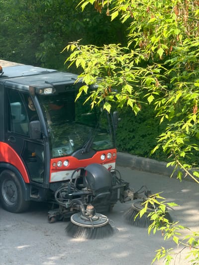 The image shows a street sweeper parked on a paved surface, likely a street or parking lot. The vehicle is primarily red and black, with a windscreen and various lights on the front. It has large circular brushes extending from the front to sweep debris. The scene appears to be outdoors during daylight hours, with abundant green foliage partially obscuring the upper and left areas of the frame. The location is stated as Tokkari, Russia, but there's no visual confirmation within the image itself to support this.