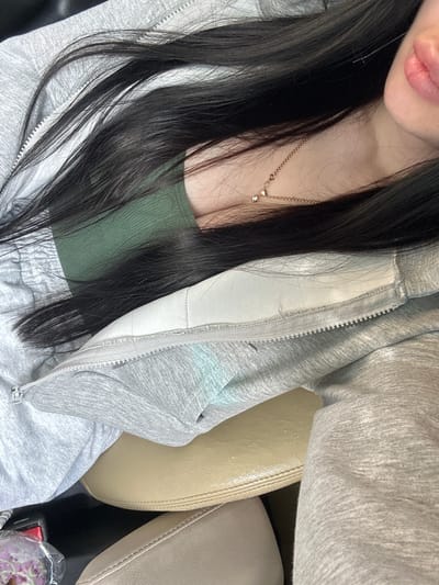 A close-up, overhead shot shows a person's upper torso and head, with dark hair cascading over a grey zip-up hoodie and a green top. A delicate gold necklace with two small diamond pendants rests on the person's collarbone. The background is out of focus, but appears to be a car interior, with a tan leather seat and a black surface visible. The image is likely taken in Monastyrshchina, Russia.