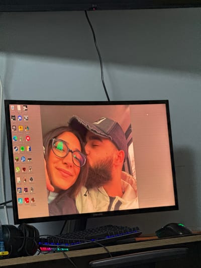 An indoor, eye-level, medium shot shows a computer monitor displaying a personal photograph of a young couple. The man is kissing the woman on the cheek. The woman wears glasses and smiles at the camera. The man wears a baseball cap. The monitor is situated on a desk, which also holds a keyboard, a mouse, and headphones. The background consists of a grey wall and a black computer cable. The photograph on the screen has a pinkish tint. The image does not provide any information about the city of Pavolche, Bulgaria.