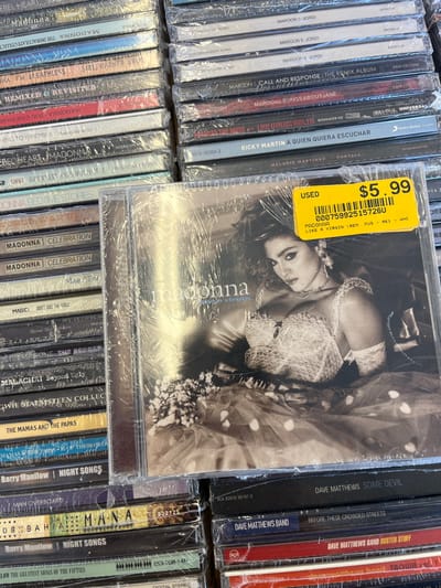 The image captures a detailed close-up of numerous compact disc (CD) albums displayed in a retail setting within the city of Edmond, United States. The CDs are primarily stacked horizontally and vertically, forming dense columns and rows. Most discs are encased in clear plastic wrap.
In the foreground and centrally positioned is a Madonna "Like a Virgin (Remix)" CD album. Its cover displays a black and white image of Madonna. A bright yellow sticker is affixed to the upper right of this album, clearly marked "USED $5.99" and includes the text "MADONNA LIKE A VIRGIN (REM MUS - RK1 - WHC)" along with a barcode "000759925157260."
Surrounding the central Madonna CD are various other albums. Discernible titles include multiple Madonna albums such as "Rebel Heart," "The Immaculate Collection," "Celebration," and "Bedtime Stories." Other visible artists and titles include "Maroon 5 - Jordi," "Maroon 5 - Call and Response," "Ricky Martin - A Quien Quiera Escuchar," "Melanie Martinez - Portals," "Barry Manilow - Night Songs," "The Mamas and the Papas," and "Dave Matthews Band - Some Devil." The environment appears to be an organized retail display for pre-owned media. No individuals are visible.