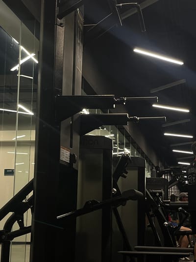 A dim, modern gym space features rows of weight machines. Linear lights accent the dark walls and equipment, creating a stark contrast. The reflective floor adds depth, mirroring the overhead lights. The machines are aligned, with visible text "MOTION" on some. People are exercising on the machines in the background.