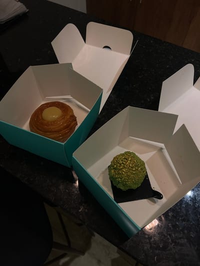 The image presents an overhead view of two open, hexagonal-shaped pastry boxes placed on a dark, speckled countertop, likely granite. Both boxes are predominantly white with a light teal or turquoise base.

The upper right box contains a distinctive green, spherical dessert with a highly textured, bubbly surface that appears to shimmer with gold accents. This item rests on a small, black, paddle-like serving piece. The lower left box holds a golden-brown, circular pastry. It features a flaky, layered exterior with a smooth, creamy yellow filling or topping in its center, resembling a fruit danish or a similar sweet baked good.

The lighting in the scene is dim, suggesting an indoor setting during the evening or night, with reflections from ambient light visible on the polished countertop. The activity depicted is the presentation or unboxing of these two unique desserts. No text is visible on the boxes or items. The context provided indicates this scene is in Mérida, Mexico.