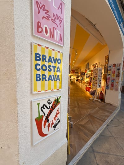 The image captures a view looking down into a shop entrance. The shop's interior is brightly lit with yellow and white striped walls. Shelves are lined with various items, including what appear to be postcards, bags, and other small souvenirs. A fire extinguisher is visible on a wall.
In the foreground, three framed artworks are mounted on a textured white wall. The first, on the left, is pink and features playful hand-drawn graphics and the text "LA BONITA." The second artwork, to its right, has yellow and white stripes and displays the words "BRAVO COSTA BRAVA" in bold blue lettering. The third piece, on the far right, has a white background with red and green illustrations of a chili pepper. It contains the Spanish phrase "Me gusta!" (I like it!) and "PICANTE" (spicy). A small signature is visible on each of these framed pieces.
Above the shop entrance, a blue banner with white text is partially visible, with "Art & Print Shop" and "TOSSA" being legible, indicating the location is likely Tossa de Mar, Spain. The overall scene suggests a lively and colorful souvenir shop, likely during daylight hours given the interior lighting.
