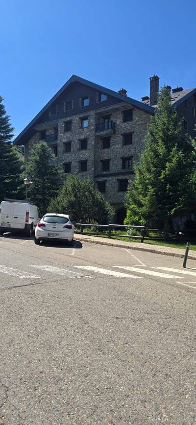 The image shows a multi-story building with a stone facade and a dark pitched roof, located in Sallent de Gállego, Spain. The building has multiple windows and balconies. In front of the building, there is a paved area with a white pedestrian crossing. Two vehicles are parked on the paved area: a white van on the left and a white car on the right. The van has a license plate that reads "3713 FTB". The car has a license plate that reads "6710 JFR". The scene is surrounded by green trees, including pine trees, and a wooden fence. The sky is clear and blue.