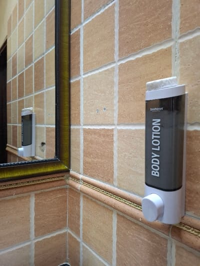 A white plastic body lotion dispenser, labeled "BODY LOTION" in black lettering and "basharani" in smaller black lettering, is mounted on a beige tiled wall. The dispenser is attached to the wall at the bottom, with a white knob in the center. The tiles are arranged in a grid pattern with grout lines in between.  The dispenser is located in a bathroom, as evidenced by the mirror in the background. The location of the bathroom is Karagita, Kenya, with coordinates -0.754, 36.437.