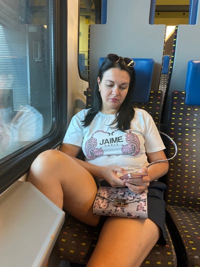A woman with dark hair is sitting on a train seat, looking down at her phone. She is wearing a white t-shirt with a pink tiger design and the text "JAIME PARIS." She has sunglasses on top of her head and a floral-patterned purse on her lap. The train appears to be in motion, as indicated by the reflection in the window. The interior of the train car is visible, with blue seats and a dark patterned fabric on the backrests. The lighting suggests it is daytime. There are no visible indications of the location being in the city of Le Brassus, Switzerland.