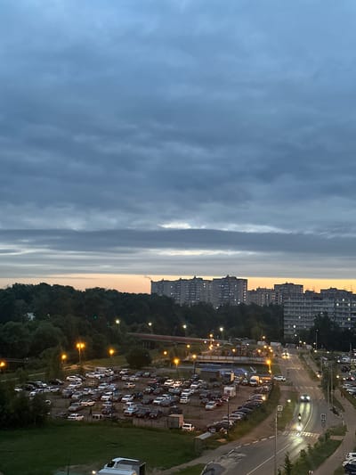 The image depicts an overcast twilight scene in Sverdlovskiy, Russia. In the foreground, a parking lot filled with numerous cars is visible, with streetlights casting a warm glow. Beyond the parking lot, a dense line of trees forms a dark green backdrop, with a red bridge arching over a dark area, possibly a river or ravine, in the middle distance. Further back, a cityscape with multi-story apartment buildings, illuminated with numerous windows, stretches across the horizon. The sky is dominated by heavy, layered gray clouds with streaks of pale orange and yellow light near the horizon, indicating the lingering twilight. A road with moving vehicles, including cars and a motorcycle, is visible on the right side of the image, bordered by sidewalks and more apartment buildings. The overall atmosphere is calm and urban, with the natural elements of trees and sky contrasting with the built environment.