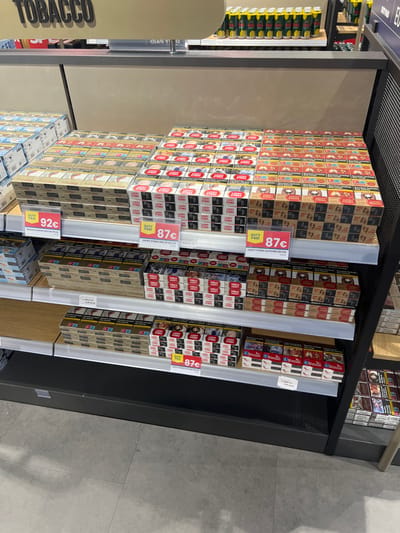 The image shows a store shelf in a supermarket located in Cologne, Germany, at the coordinates 50.879, 7.121. The shelf is stocked with cigarette packs, arranged in rows and columns, with price tags attached to each row. There are three rows of cigarette packs on the shelf. The top row is filled with packs of cigarettes marked with red and white coloring, while the bottom two rows have cigarette packs marked with blue and white coloring. The image captures a view of the shelf from an angle, revealing the interior of the store behind the shelf. The store environment is brightly lit and has a light-grey tiled floor. There are no people visible in the image.