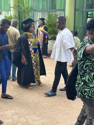 The image displays an outdoor scene in Madina, Ghana, featuring a gathering of individuals on a light brown paved area composed of interlocking rectangular pavers. A modern building with large, reflective green-framed windows and light green architectural accents forms the background.
In the foreground, from left to right:
A male with dark skin, wearing a blue long-sleeved shirt, blue trousers, and dark slip-on sandals, stands looking down at a horizontally held smartphone.
Adjacent to him, a female with dark skin, wearing a black graduation gown over a vibrant multi-colored dress featuring yellow, blue, and black patterns, dons a yellow stole and a black mortarboard cap. She holds a dark rectangular object and a smartphone in her left hand and wears yellow open-toed high-heeled sandals. She looks slightly to her right with a neutral expression.
Next to her, a male with dark skin, wearing a white traditional Ghanaian smock with a gathered mid-section and black trousers, stands facing left, looking directly towards the female graduate. He wears dark blue dress shoes.
Partially visible on the far right foreground is the right side of a person wearing a black and bright green abstract patterned shirt, dark trousers, and dark shoes.
In the mid-ground, positioned on a raised platform closer to the building, a second female with dark skin wears a black graduation gown, a gold and purple stole, and a black mortarboard cap over a light peach-colored garment. She looks slightly to her left with a faint smile.
Further in the background, near the left, a partially obscured male with a bald head wears a light-colored top and light blue jeans, appearing to hold a camera. Green potted plants, resembling palm trees, are visible near the building's facade. Other indistinct figures are visible through the building's windows. The scene is illuminated by bright natural daylight.