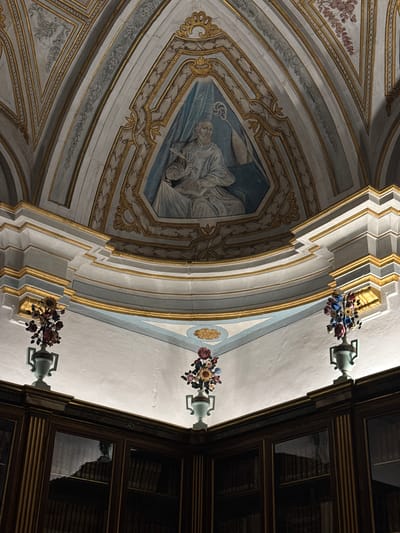 This media file depicts the interior of a grand, ornate building, possibly a church or a library, with a focus on the ceiling architecture and decorative elements. The ceiling is a complex structure of curves and recessed panels, adorned with gold trim and intricate painted designs. A significant fresco occupies a triangular recess in the ceiling, depicting a figure in repose, possibly in a religious or historical context. Surrounding the fresco are elaborate patterns and ornamental borders.
Attached to the ceiling are decorative light fixtures, resembling urns, from which cascades of artificial flowers hang. These floral arrangements add vibrant pops of color against the muted tones of the architecture. To the right of the image, a large, dark wooden structure with multiple glass panes dominates the frame. These panes reflect the interior of the building, hinting at the presence of bookshelves filled with books, suggesting a library setting. The lighting suggests it is daytime, with sunlight casting dramatic shadows, highlighting the architectural details and creating a sense of depth. The overall impression is one of historical grandeur and artistic richness, conveying a solemn and awe-inspiring atmosphere. No visible text is present in the image.