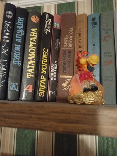 The image depicts a close-up, static indoor scene set on a wooden shelf, located in Baranavichy, Belarus. The primary subjects are a row of ten books, predominantly with Cyrillic titles, and a singular decorative figurine.
From left to right, the visible book spines are:
1. A dark blue/black spine, titled "ЭРНЕСТ ХЕМИНГУЭЙ" (Ernest Hemingway) in white Cyrillic, with a circular emblem containing a cross at the top.
2. A dark blue/black spine, titled "ДЖОН АПДАЙК" (John Updike) in light blue Cyrillic, marked with a stylized 'K' logo.
3. A dark blue/black spine, titled "ФАТА-МОРГАНА" (Fata Morgana) in yellow Cyrillic, featuring a red number '4' and an orange yin-yang-like symbol at the top.
4. A dark blue/black spine, titled "ЭДГАР УОЛЛЕС" (Edgar Wallace) in yellow Cyrillic, with "АНГЛИЙСКИЙ БОЕВИК" (English Thriller) printed above it.
5. A reddish-pink spine, displaying the number "20", with smaller text "Дон Пендлтон" (Don Pendleton) and a "Met" logo.
6. A brown spine, titled "ЭРИХ МАРИЯ РЕМАРК" (Erich Maria Remarque), "ТЕНИ В РАЮ" (Shadows in Paradise), and "ЖИЗНЬ ВЗАЙМЫ" (Borrowed Life) in white Cyrillic, stacked vertically.
7. A light brown spine, titled "Жорж Санд" (George Sand) in white Cyrillic.
8. A light blue spine featuring a graphic resembling Mickey Mouse ears.
9. A grey-green spine, titled "Харпер Ли" (Harper Lee) in white Cyrillic.
10. A grey-green spine, titled "Джон Болл" (John Ball) in white Cyrillic.
Positioned in front of the sixth, seventh, and eighth books is a glossy, multi-colored figurine of a cartoonish horse or pony. The figurine has a yellow head, a red torso, orange legs, and black hooves, depicted in a seated posture. It rests on a textured, golden base, which appears to represent a pile of coins or treasure.
The books and figurine are situated on a dark wooden shelf. The background consists of a wall covered in vertically striped wallpaper, with alternating bands of dark green, light green, and beige. Illumination originates from the front, causing reflections on some book spines and the figurine.