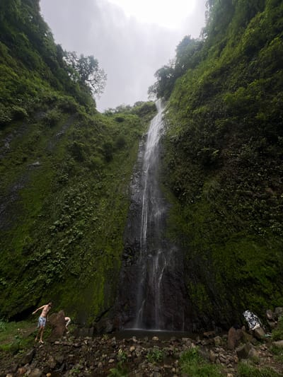 The image depicts a scenic view of a waterfall cascading down a steep, moss-covered cliff face. The surroundings are lush with vegetation, with trees growing on the upper ledges of the cliff. The sky is overcast, suggesting a cloudy day.
In the foreground, near the base of the waterfall, two individuals are present. One person, wearing patterned swim trunks, stands on a rocky outcrop near the water's edge, appearing to be posing. Another person is partially visible, bent over and interacting with the rocks and water. Various rocks and greenery are scattered around the area. A patterned garment is draped over a rock to the right of the scene.
The location is identified as the city of Altagracia, Nicaragua, implying this is a natural attraction within or near that city.