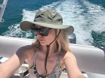 A woman wearing a swimsuit and a khaki sun hat with sunglasses perched on top is on a boat traveling through water. The woman's head is oriented downwards in the image frame. The water is visible in the background, displaying visible ripples and splashes indicative of the boat's movement. The scene is set in Unknown, Mexico.