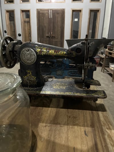 An antique sewing machine sits on a wooden table in what appears to be a covered outdoor or semi-open space. The sewing machine is black with intricate gold detailing, showing signs of age and wear. Behind the table, a building with large wooden doors and several windows is visible. The overall setting suggests a rustic or traditional environment, potentially in Tambakreja, Indonesia, given the architectural style. There are no people present in the image. A partially visible glass jar is in the foreground, to the left of the sewing machine. The lighting is dim, casting shadows and highlighting the texture of the objects.