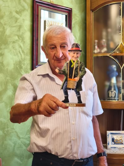 In Campochiaro, Italy, an elderly Caucasian man with white hair and fair skin stands in what appears to be a home interior. He is wearing a light-colored, vertically striped short-sleeved button-down shirt and dark trousers. The man is holding a small, figurine of a person dressed in a green coat, a black hat with an orange band, and a green scarf. The figurine is standing on a wooden base with the name "NINO" inscribed on it. The background shows a textured green wall on the left and a wooden cabinet with glass doors on the right. Inside the cabinet, several bottles, one prominently displaying the number "80," are visible, along with a framed picture of two rings. The man's right hand is extended, holding the figurine.
