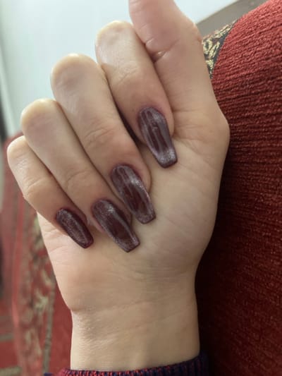 A hand rests against a background of reddish-brown fabric and a patterned surface. The skin is fair, with visible veins and creases. The nails are painted a dark reddish-brown color with a shiny texture, shaped in a coffin style. The fingers are slightly curled, suggesting a relaxed posture. The focus is on the hand and its manicured nails.