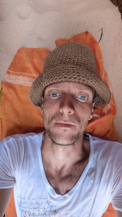 A light-skinned male with blue eyes, a short beard, and a brown knit cap lies on an orange patterned cloth. The cloth rests on sandy ground. He wears a white t-shirt with designs on the front. The scene is outdoors. The location is Phuoc Dinh, Vietnam.