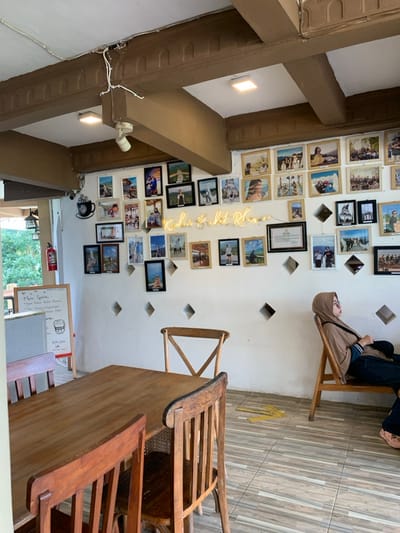 A wooden table and four chairs are arranged in a corner of a cafe located in Kecamatan Borobudur, Indonesia at -7.606 latitude and 110.18 longitude. The walls are white and adorned with framed photographs, including a picture of the iconic Borobudur Temple. A neon sign that reads "Kedai & Restoran" is mounted above the photographs. A woman in a brown hijab sits on a wooden chair with a tan cushion. Her legs are crossed and she is looking down at her phone. The wooden floors have a tiled pattern. A fire extinguisher is visible in the corner of the room, and a menu board is leaning against the wall. There is a large window in the background that looks out onto a verdant garden. The cafe is brightly lit and has a relaxed ambiance.