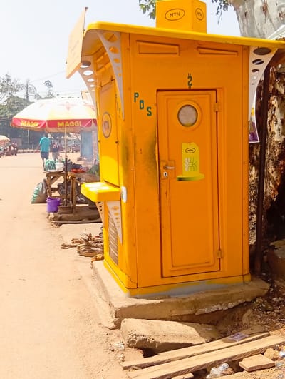 This is a medium shot, eye-level, outdoor, daytime photograph taken in Barikin Gidar Kanat, Nigeria. The primary subject is a bright yellow MTN mobile money kiosk situated on a concrete base. The kiosk has the letters "POS" printed vertically on its side and a circular window above a locked door. A SIM card graphic is affixed to the door. Above the kiosk, a yellow MTN branded sign is visible.
To the left of the kiosk, a street market scene unfolds. Two umbrellas with "MAGGI" branding are set up. Underneath them, a wooden table displays produce, including what appears to be tomatoes and green vegetables, alongside other market goods like a purple bucket and a green sack. Two men are visible near the table; one wearing a blue shirt and the other a dark t-shirt with graphics. In the background, the street is visible with a few vehicles and other market stalls. The ground is a mix of dirt and gravel. A large tree trunk with rough bark is on the right side of the frame, partially obscuring the kiosk. Wooden planks and debris are scattered on the ground in the foreground. The sky is bright and clear.