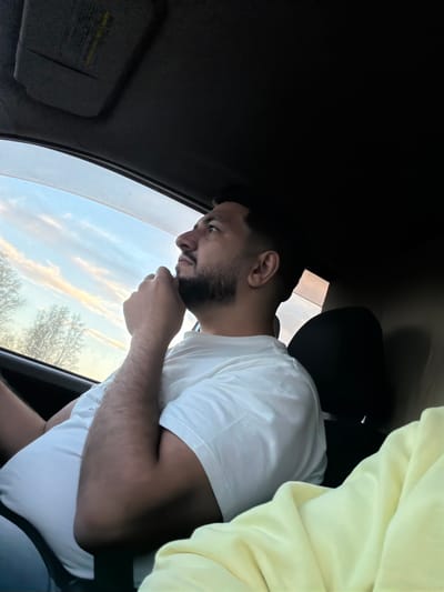 A medium shot from inside a vehicle facing a man in a white T-shirt who is looking out the window. He has dark hair and a beard, and he is touching his chin. Outside the window, the sky is visible, and there are trees in the distance. A light yellow cloth is draped over the passenger seat and part of the man’s shoulder. The sun visor is down on the driver's side, and the car interior is mostly dark. The location and city in Russia cannot be determined from this image.