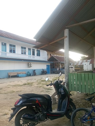 This outdoor image, taken in Dayahtuha, Indonesia, captures a bright daylight scene featuring vehicles and buildings. The perspective is from underneath a covered structure with a corrugated metal roof and wooden supports, visible in the upper and left parts of the frame.
In the immediate foreground on the right, a black scooter is parked on a light-colored, dusty ground. The word "SCOOP" is distinctly visible on its side, identifying it as a Honda Scoopy. Partially visible behind it is a blue bicycle. To the right, a green wooden fence or barrier runs across the mid-ground.
The central and left portion of the image is dominated by a multi-story building featuring white walls with a blue base and blue-framed windows. A white balustrade decorates a balcony on its upper level. An air conditioning unit is mounted on the building's exterior. Outside this building, a wooden bench and a green plastic chair are visible, and a person can be seen sitting on another scooter or motorcycle. In the background, other structures with corrugated roofs and a white van or truck are faintly discernible.
The bright sunlight, particularly from the left side of the frame, suggests a clear and sunny day, likely midday or early afternoon, casting strong highlights and shadows across the scene.