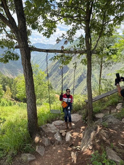 A person sits on a swing suspended between two trees, in an outdoor setting with mountains visible in the background. The swing is made of metal and chains. The person wears sunglasses, a red and white jacket, blue jeans, and brown boots, and has a small orange bag on their side. The trees are mature with thick trunks and dense foliage, partially obscuring the view. The ground is covered with grass, rocks, and soil. Another person's hands holding a cell phone are visible in the upper left corner, presumably taking the photo. The landscape suggests the location is likely in a mountainous region, as indicated by the Lũng Cú Commune, Vietnam description.