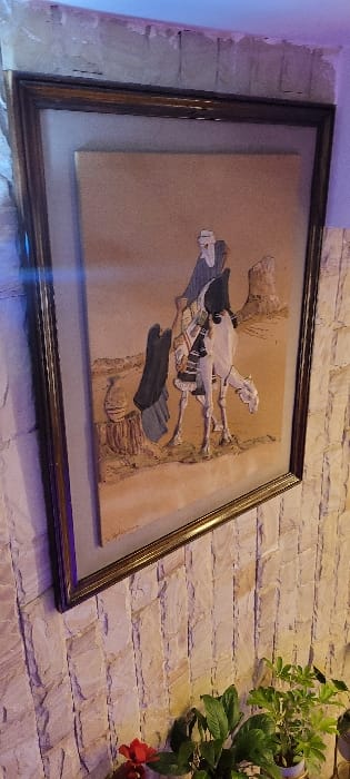 A framed artwork hangs on a textured, light-colored brick wall. The art depicts a scene with figures, possibly a horse and rider, in a stylized or abstract manner. The artwork features raised elements, creating a three-dimensional effect. To the right of the artwork, a potted green plant with some red flowers adds a touch of nature. The wall is illuminated with soft, diffused light.