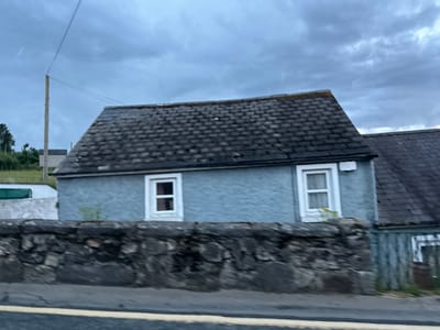 The image depicts a slightly blurred, horizontal view of a modest, single-story house in Rathvilly, Ireland. The house, painted a pale blue, has a dark, sloped roof covered in what appears to be slate tiles. Two white-framed windows with curtains are visible on the front of the house. In front of the house is a low, rough-hewn stone wall. Beyond the wall, to the left of the house, is a grassy slope with some trees and a utility pole with wires. To the right of the blue house, part of another building with a similar dark roof and faded blue siding is visible. The sky above is overcast with heavy, dark clouds. A yellow line is faintly visible on the road in the foreground, suggesting the image was taken from a vehicle in motion. The overall scene suggests a somewhat weathered and rural setting under a brooding sky.