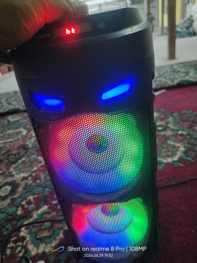 The image shows a close-up of a black portable speaker with blue LED lights resembling eyes and a colorful light show on its mesh speaker grills. A hand is visible in the upper left corner, partially obscuring a red "bt" indicator light and some serial numbers. The speaker is placed on a patterned rug, and the background suggests an indoor or semi-outdoor setting with blurry figures and structures visible. A watermark in the lower right corner indicates the photo was taken with a "realme 8 Pro | 108MP" on "2026.06.29 19:52". The presence of the specific watermark and date format strongly suggests this image is not from Bokhtar, Tajikistan, or the current time.