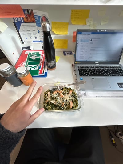 The image depicts an indoor office or workspace in Illkirch-Graffenstaden, France. A person's left hand, covered by a dark, textured sleeve, is positioned on a white desk surface, hovering above a rectangular clear glass container holding a meal. The meal consists of green spiral pasta mixed with dark leafy greens, topped with flakes of pink salmon and grated white cheese, with a silver fork resting within.

To the left of the food container are two supplement bottles with white caps: one dark grey with "MY[FITNESS/FUEL]" text, and one amber-colored with "VITAMIN B" visible. Adjacent to these is a black insulated water bottle with a silver cap and a brown base, featuring a white mountain-range design. Further left, a white upright file holder contains papers and a partially visible blue and white poster. A red folder labeled "REGISTR[ER/E]" and featuring green emergency exit signs is stacked on the desk in front of the file holder.

On the right side of the desk, an open silver Acer laptop displays an email client interface on its screen. Stickers indicating "1080p Full HD" and "Intel Core i5" are present on the laptop's palm rest. The white wall or partition behind the desk is adorned with numerous yellow and orange sticky notes containing handwritten text. Below the desk, the person's legs, clothed in dark trousers, are visible, along with a black power cable and a partially visible white power strip with a red switch.