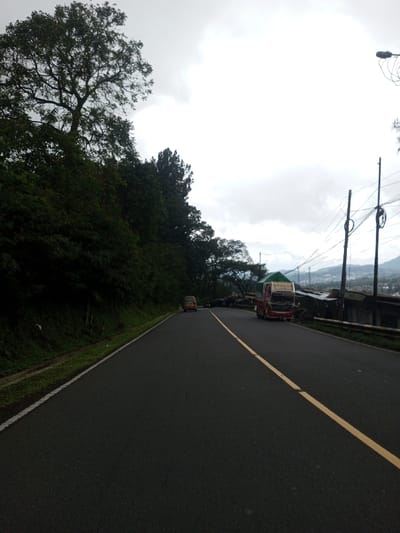 The image presents a two-lane asphalt road in Deothang, Bhutan, under an overcast sky. The road features a solid white line marking its left edge and a dashed yellow line separating the lanes, extending into the distance with a slight right curve. To the left, a steep embankment is densely covered with dark green trees and lush vegetation. On the right side of the road, a red truck with a green tarpaulin cover over its bed is parked, facing away from the camera. Further down the road, a light-colored van or minibus is also visible. Adjacent to the parked vehicles, a row of low-rise structures, possibly market stalls, with corrugated metal roofs are present. Two dark utility poles with multiple overhead power lines stand on the right, alongside a metal guardrail. In the distant background on the right, hills and a scattered settlement are discernible beneath the cloudy conditions. No people are visible.